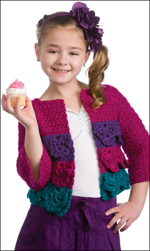 Child wearing a crochet cardigan sweater with layers of lacy ruffles and sparkly yarn. Sweater is pink, purple, and teal. She is also wearing a white shirt and purple skirt, and holding a cupcake.